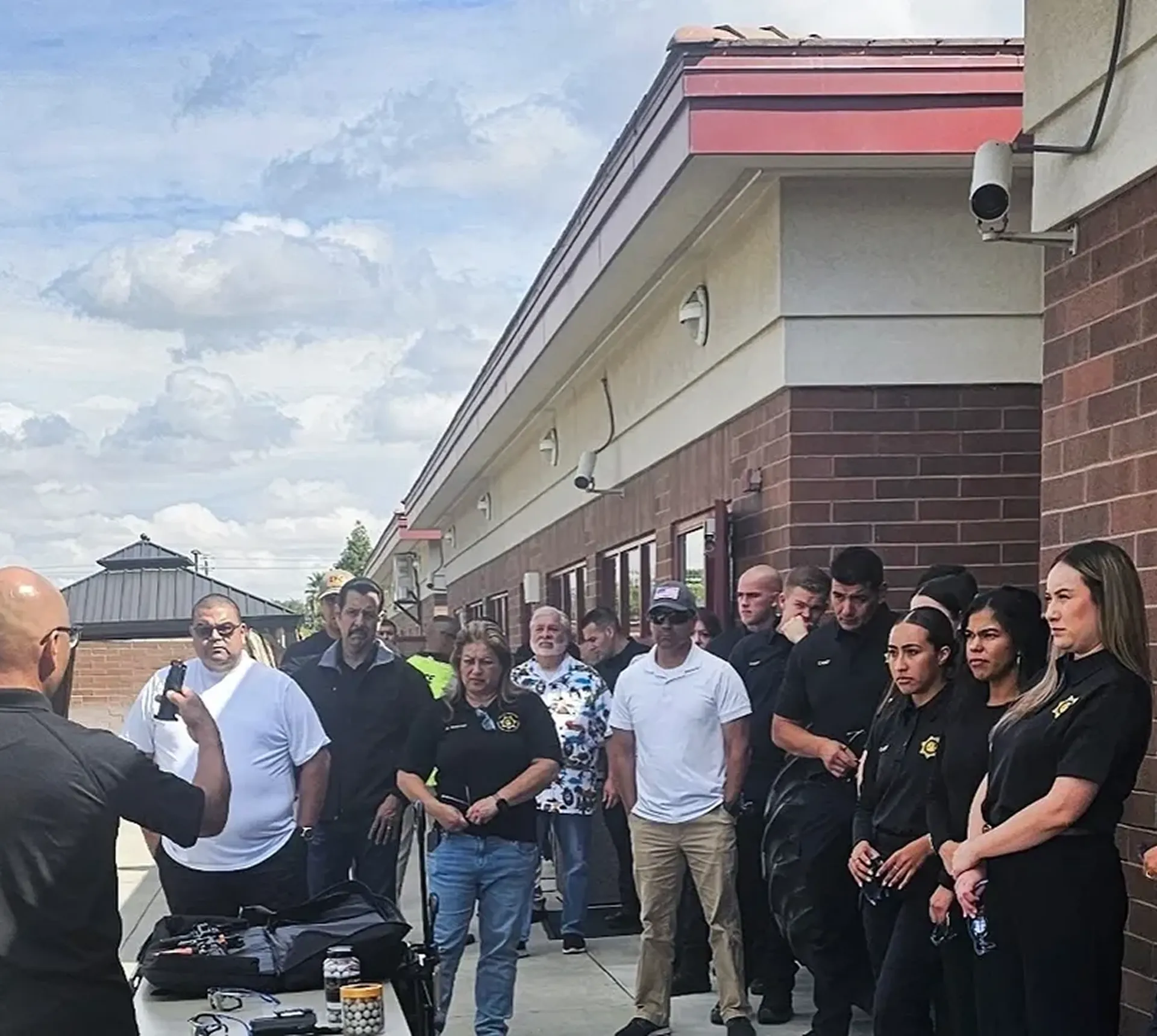 Group gathered outside brick building, listening attentively.